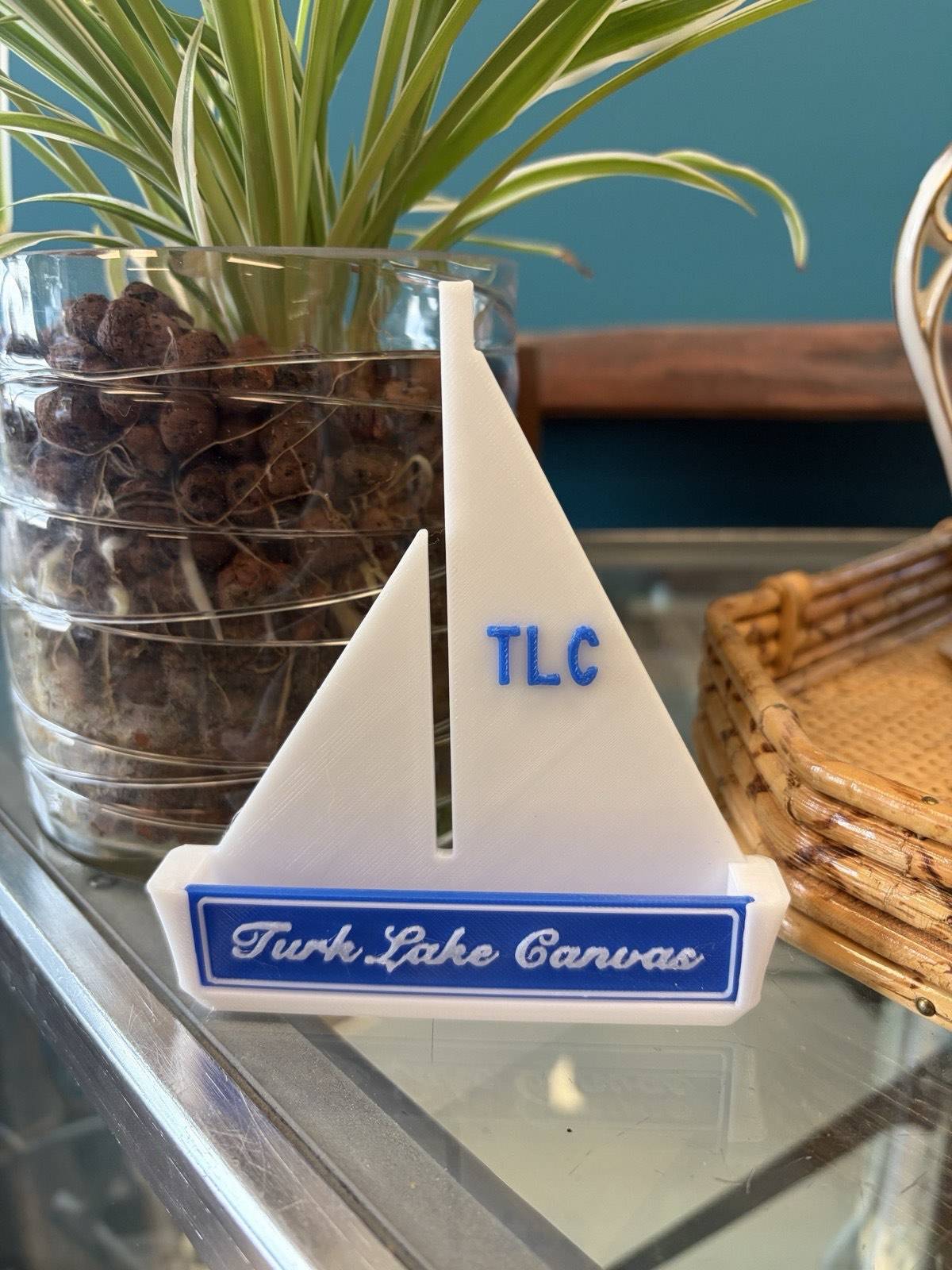 A custom business card holder shaped like a sailboat, with TLC on the sail and Turk Lake Canvas in script on the hull