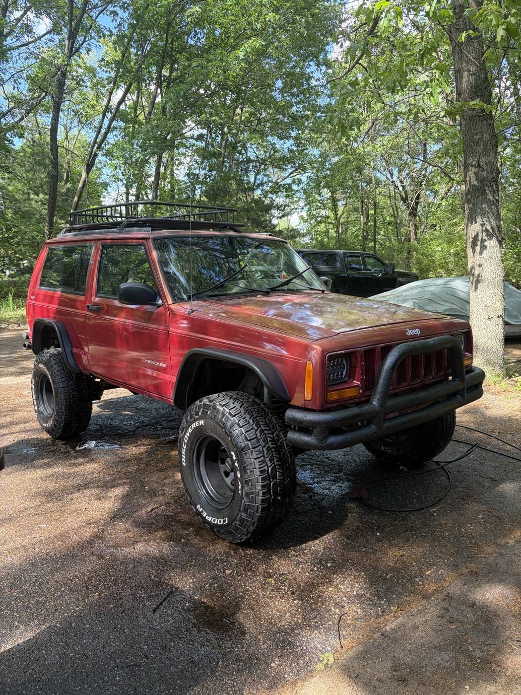 The 1998 Jeep Cherokee XJ in red, lifted on large tires with a front bumper and a roof rack, parked in the trees
